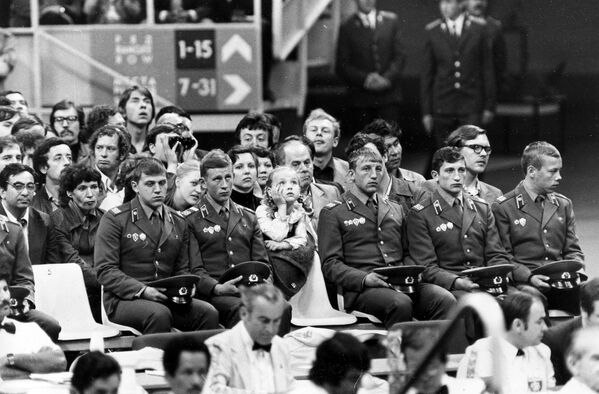 Маленькая девочка кладет голову на руку, наблюдая за боксерским матчем во время XXII летних Олимпийских игр в Москве. Советские солдаты, сидящие перед маленькой болельщицей, обеспечивают общие меры безопасности во время игры. - Sputnik Беларусь