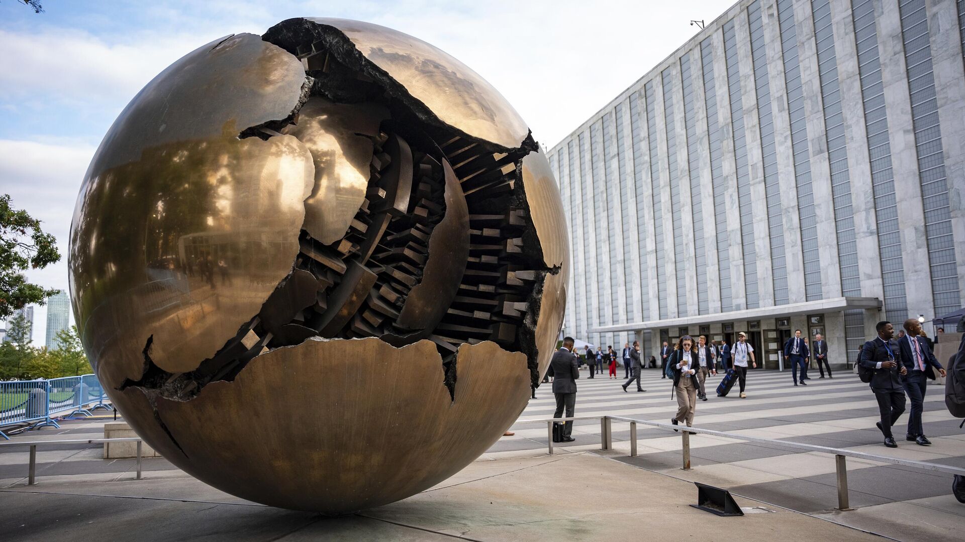Люди прогуливаются по площади возле скульптуры Сфера внутри сферы перед штаб-квартирой ООН