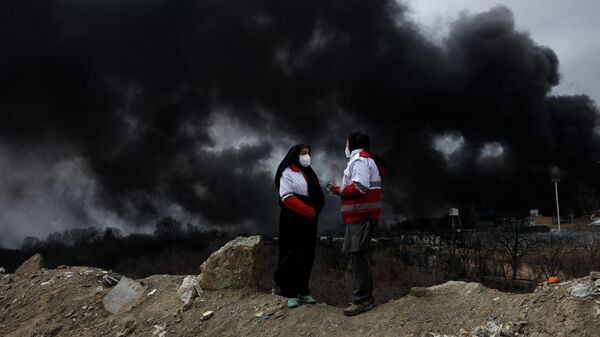 Члены Красного Полумесяца разговаривают друг с другом на фоне поднимающегося дыма после сообщения об ударе по топливным резервуарам в Шахране, Иран - Sputnik Беларусь