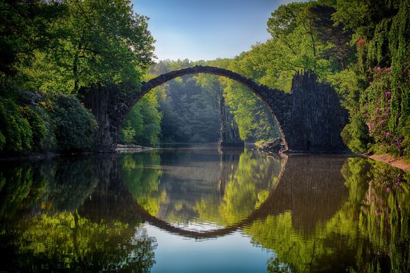 Чертов мост (Rakotzbrücke) — это каменная арка в парке Кромлау в 120 км от немецкого Дрездена, построенная в 1860 году. Базальтовые камни, из которых сделан мост, доставлялись сюда из разных областей Саксонской Швейцарии и Богемии. Вместе с отражением в озере Чертов мост образует круг. Этот факт породил легенды об архитекторе, заключившем сделку с дьяволом, и о том, что здесь находится портал в другие миры. - Sputnik Беларусь