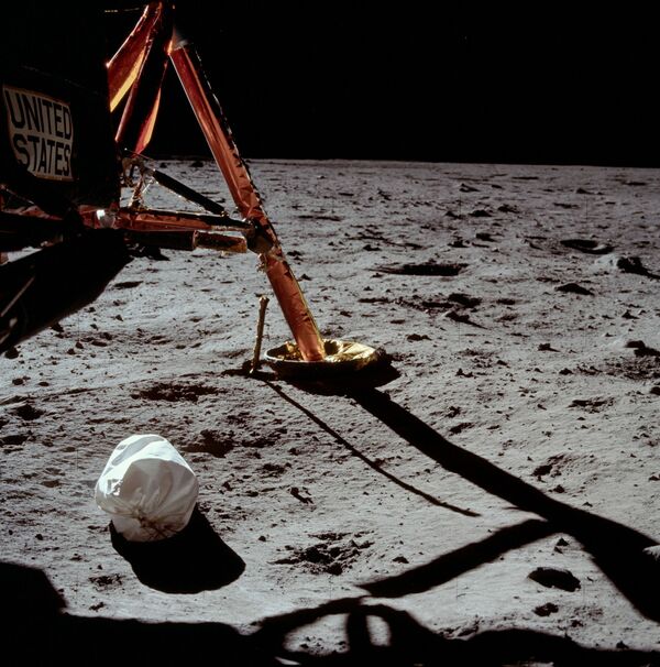 Первая фотография, сделанная Армстронгом после выхода на поверхность Луны. После открытия выходного люка он начал потихоньку в него протискиваться спиной. Выбравшись на площадку над лестницей, Армстронг первым делом отрепетировал возвращение в лунный модуль. Затем, взяв мешок с мусором, который ему передал Олдрин, он снова выбрался на площадку и выбросил мешок на лунную поверхность. Белый пакет на переднем плане — мешок с мусором.  - Sputnik Беларусь